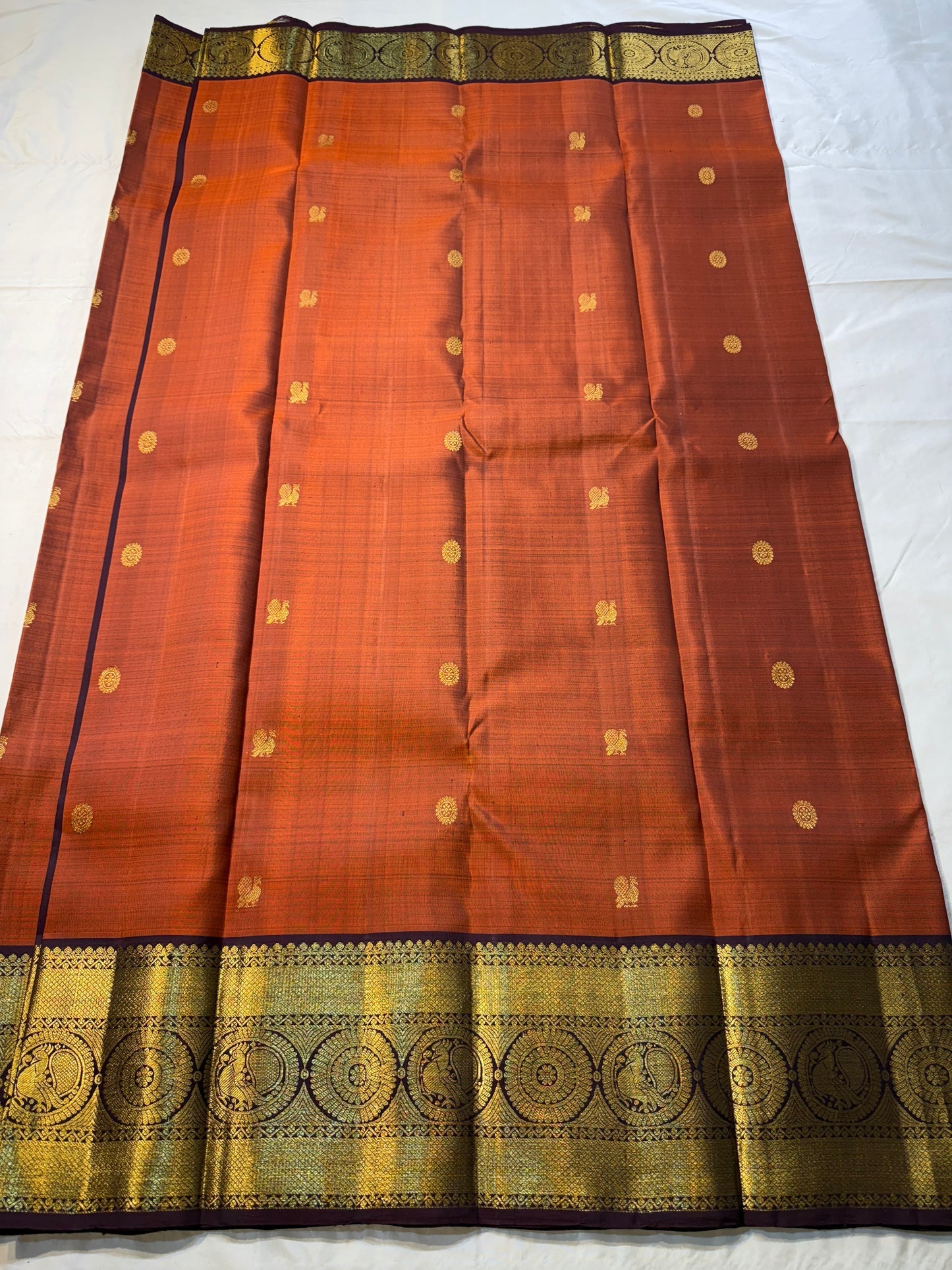 Rust Orange Supreme – Korvai Kanchipuram Pure Silk Saree
Shot rust-orange body with gold chakram & mankolam buttas all over
Deep purple korvai border with annam & chakram zari medallions
Rich vadamalli zari-brocade pallu | Contrast blouse | Fall & Pico Do