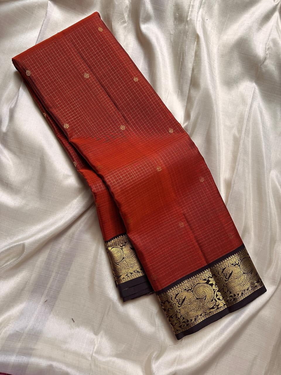 Mantra – Rust brown with coffee brown Kanchipuram Silk Saree with zari Checks with small buttas all over and Antique Peacock zari Border showcasing regal peacocks and floral vines. A richly detailed pallu completes this heritage Zari kattam