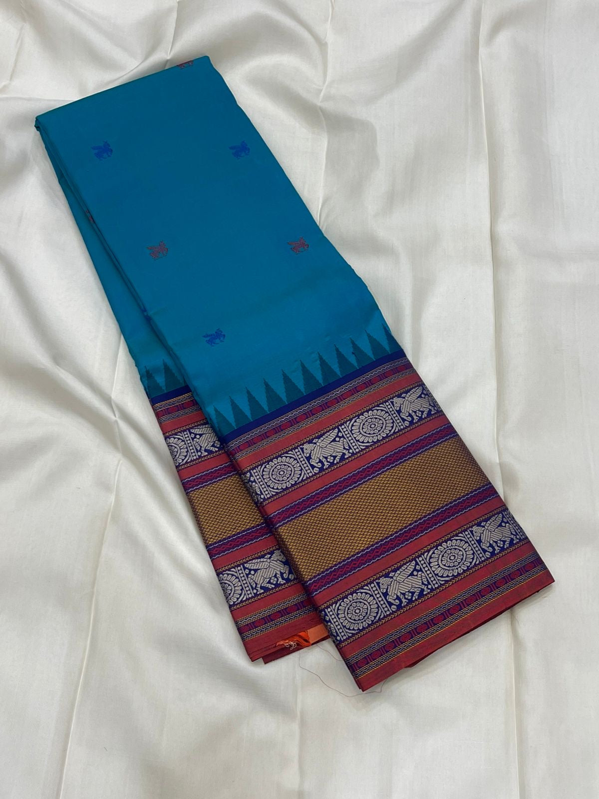 Peacock-teal body with woven silk-thread buttas in navy & coral hues
Korvai contrast ruby-maroon borders with gaja-chakram & temple motifs
Grand varisaipettu striped pallu in navy, mustard & maroon tones
Silk Mark Certified • Ships in Two Weeks