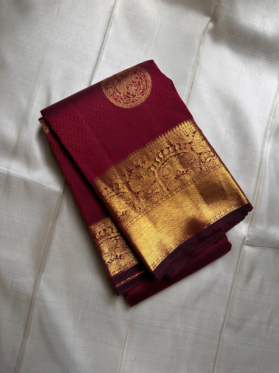 A majestic maroon Kanchipuram silk saree, adorned with intricate golden zari featuring the mythicalYazhi (Yali ) motifs—a symbol of divine strength and protection in Hindu mythology. The grand temple-style gold border adds depth and regality, making this