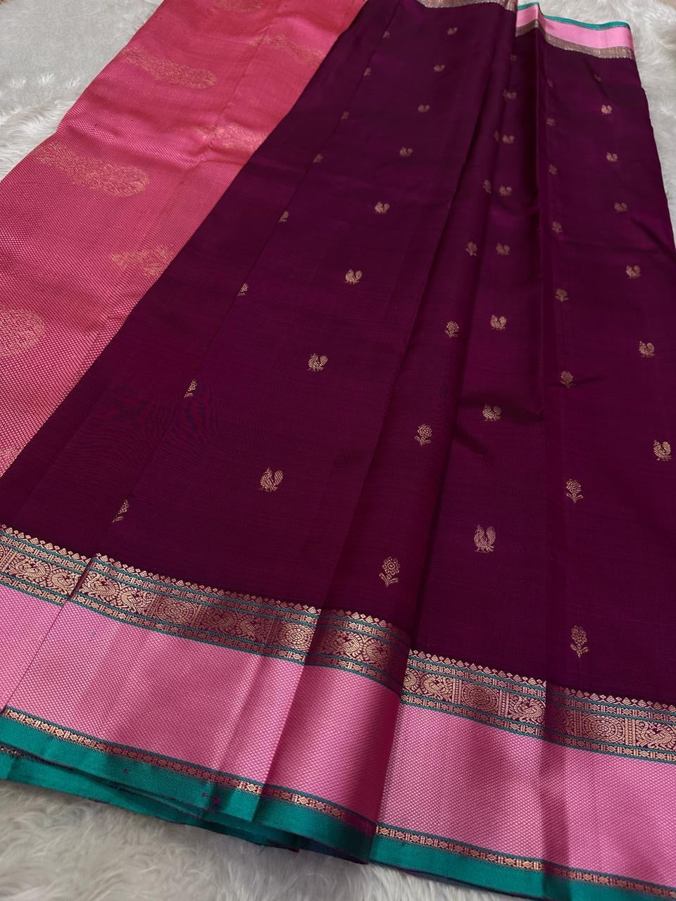 A regal magenta silk with intricate zari buttas scattered delicately across the body. The border is a stunning blend of soft pink and teal, featuring fine gold silver zari weaving and temple motifs