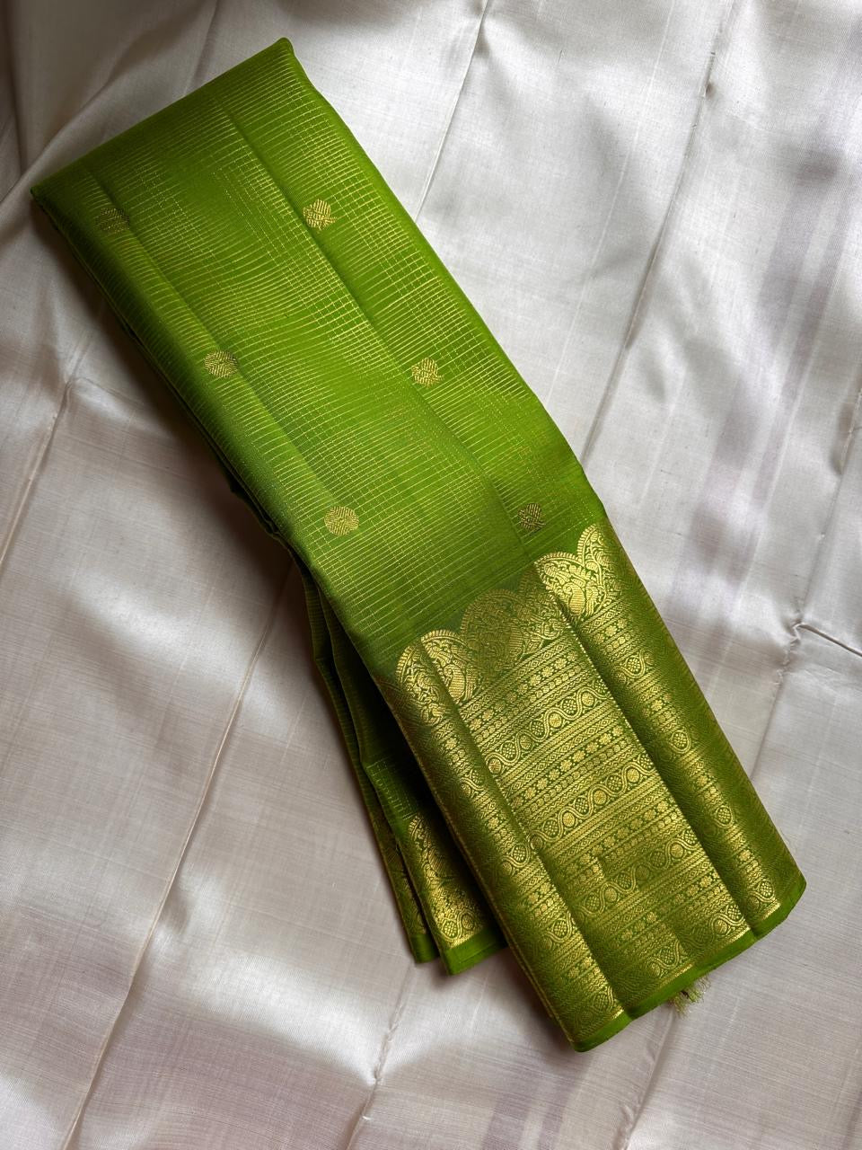 A radiant parrot green Kanchipuram silk saree that dazzles. The body features a delicate grid-like zari weave with small gold buttas scattered. The rich gold zari pallu stands out with grand temple-inspired arches and intricate motifs.