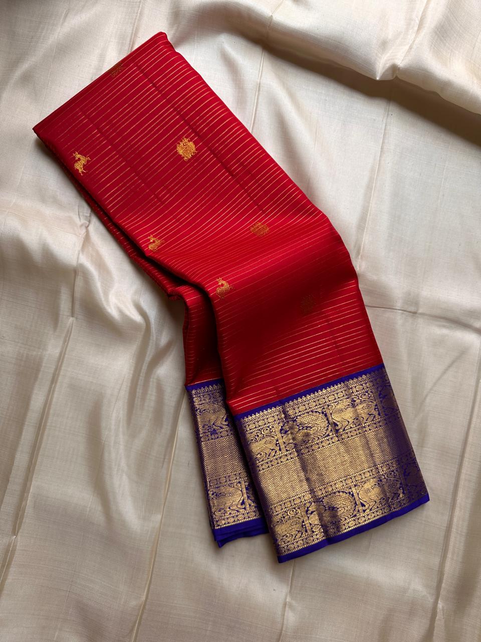 Raaga – Red and Blue Pure Kanchipuram Silk Saree with Gold Zari Border
Features a striped red body with small gold buttas and a royal blue pallu.
Finished with intricately woven peacock motifs in pure zari across the border.