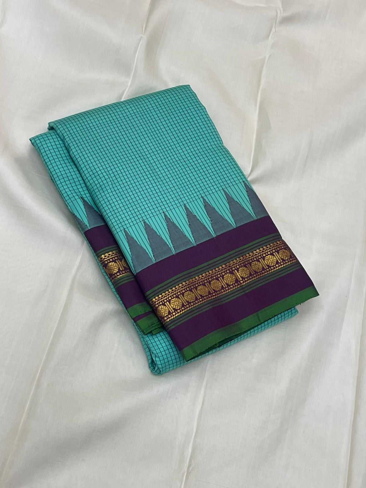 Gopura Neelam — Kanchipuram Pure Silk Saree
 
 
Teal-green body with micro kattam checks for a sleek drape
Regal aubergine korvai border with gopuram/temple motifs & gold zari
Parrot-green selvedge for a fresh, festive pop of contrast
Aubergine pall