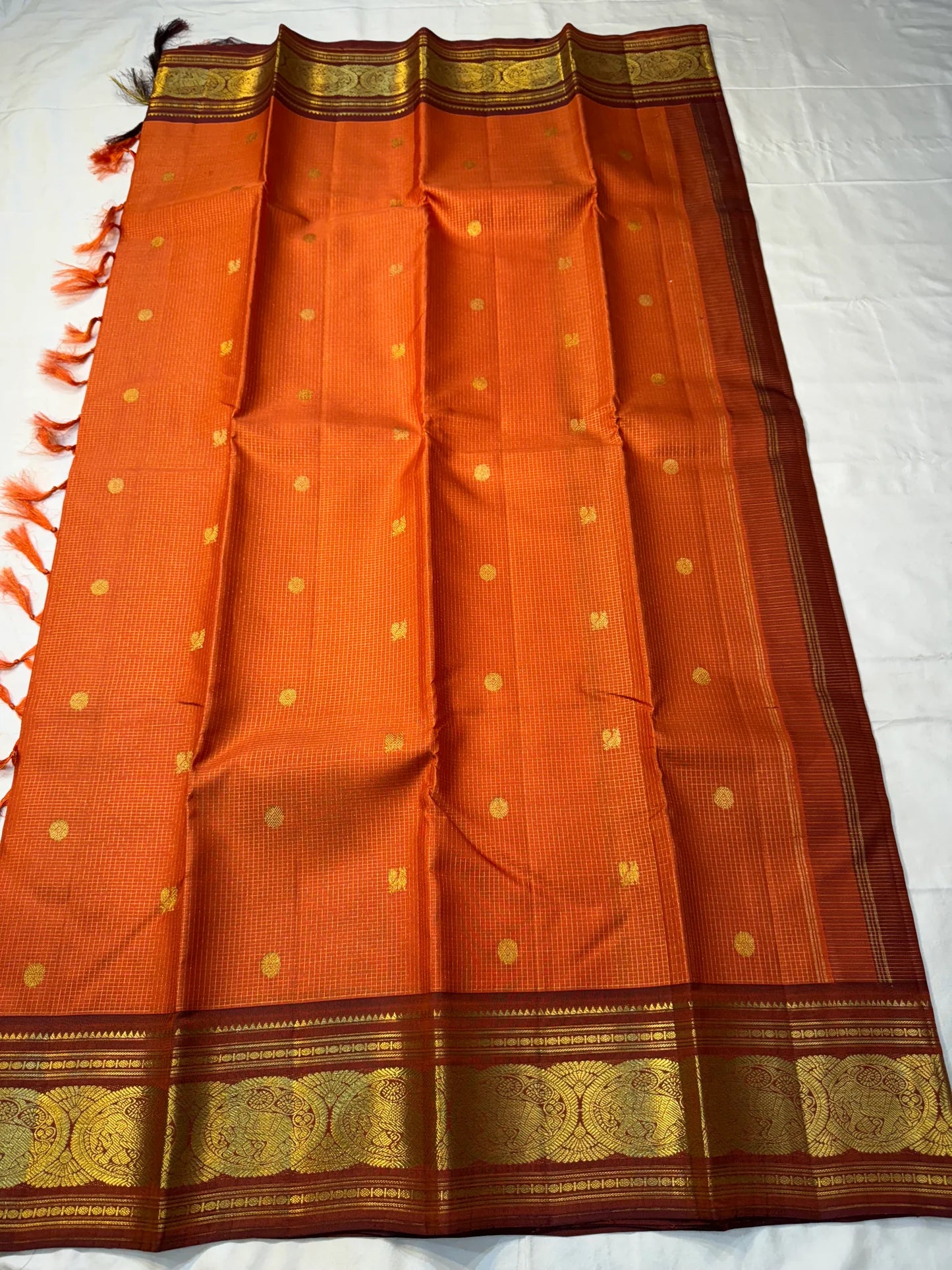 TEJAS – Vibrant Rust Orange Podi Kattam Kanchipuram Silk Saree
Micro-checks body with scattered gold chakram buttas; rich contrast maroon korvai borders
Grand circular annapakshi medallions in lustrous zari; ornate-jaal pallu & tassels
Silk Mark