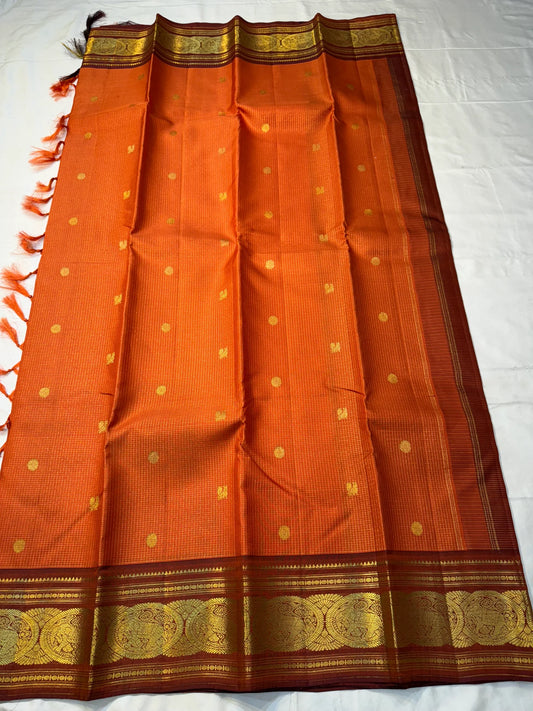 TEJAS – Vibrant Rust Orange Podi Kattam Kanchipuram Silk Saree
Micro-checks body with scattered gold chakram buttas; rich contrast maroon korvai borders
Grand circular annapakshi medallions in lustrous zari; ornate-jaal pallu & tassels
Silk Mark