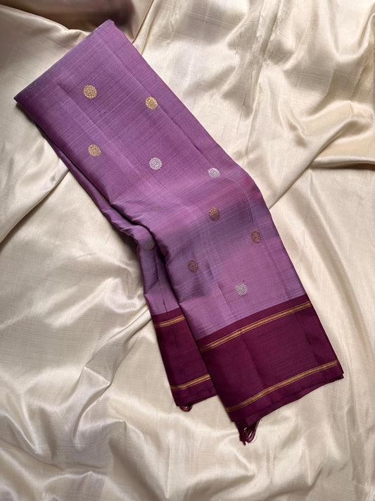 Ilam  Vadamalli Chakram — Kanchipuram Pure Silk
Pastel lilac–mauve body with gold & silver chakram buttas
Deep plum contrast border with twin kaddi zari lines
Plum pallu with elegant vertical zari stripes; lilac tassels
Silk Mark Certified •