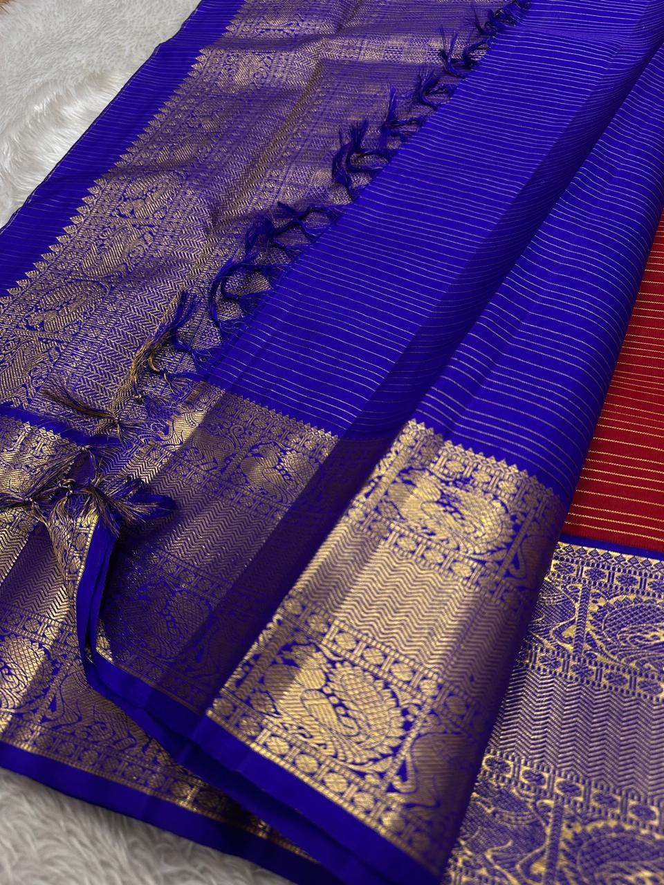 Raaga – Red and Blue Pure Kanchipuram Silk Saree with Gold Zari Border
Features a striped red body with small gold buttas and a royal blue pallu.
Finished with intricately woven peacock motifs in pure zari across the border.