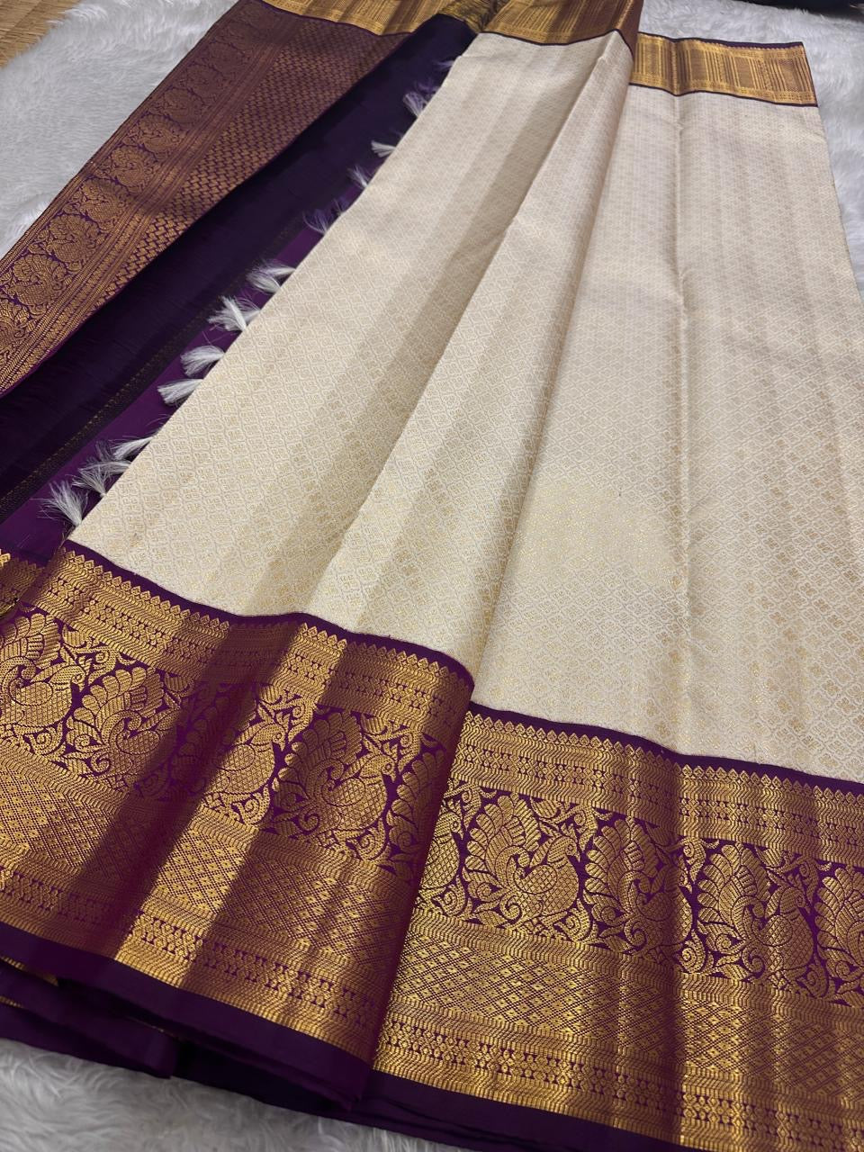 Ivory silk meets tradition in this elegant Kanchipuram saree. With a deep purple border and Mayuri motifs, it’s perfect for moments that call for quiet richness.