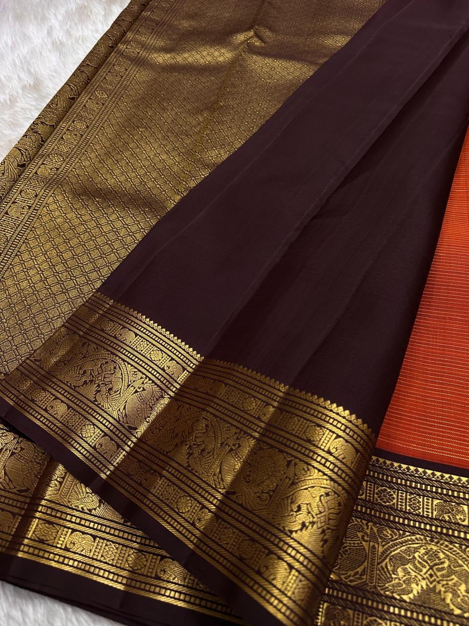 Kavi — oosi kattam Pure Kanchipuram Silk Saree
Burnt Orange (saffron–rust) body
Deep coffee-maroon contrast border with grand antique-gold zari
Rich brocade pallu; peacock & floral vines in the border
Silk Mark Certified