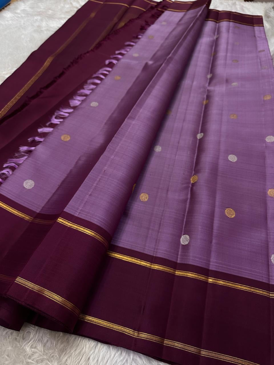 Ilam  Vadamalli Chakram — Kanchipuram Pure Silk
Pastel lilac–mauve body with gold & silver chakram buttas
Deep plum contrast border with twin kaddi zari lines
Plum pallu with elegant vertical zari stripes; lilac tassels
Silk Mark Certified •