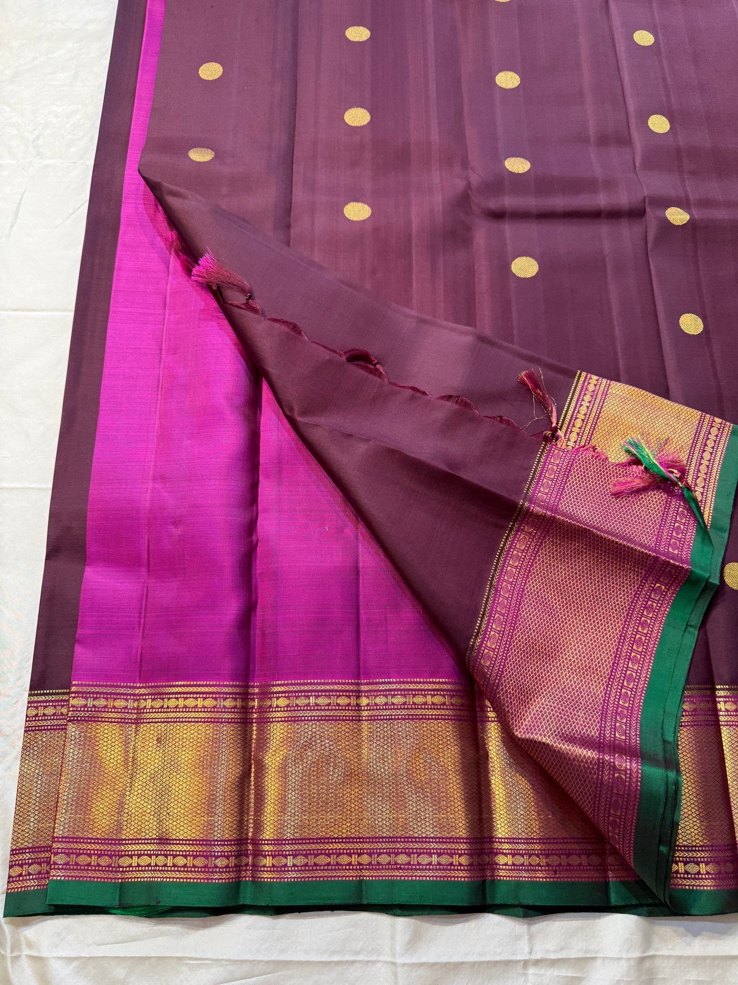 Plum & Magenta Round Butta – Pure Kanchipuram Silk Saree
Deep plum body with bold round gold zari buttas
Contrast green selvedge & gold tissue borders with magenta detailing
Grand magenta-pink pallu with full zari diamond jaal & geometric panels
Silk Mark