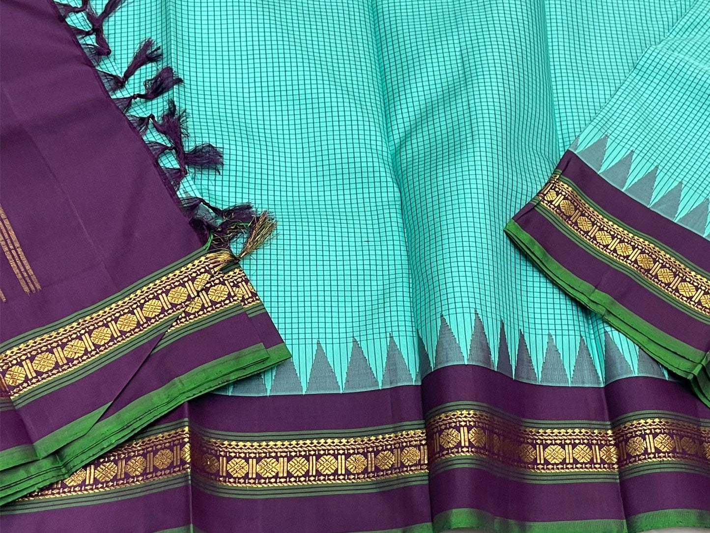Gopura Neelam — Kanchipuram Pure Silk Saree
 
 
Teal-green body with micro kattam checks for a sleek drape
Regal aubergine korvai border with gopuram/temple motifs & gold zari
Parrot-green selvedge for a fresh, festive pop of contrast
Aubergine pall