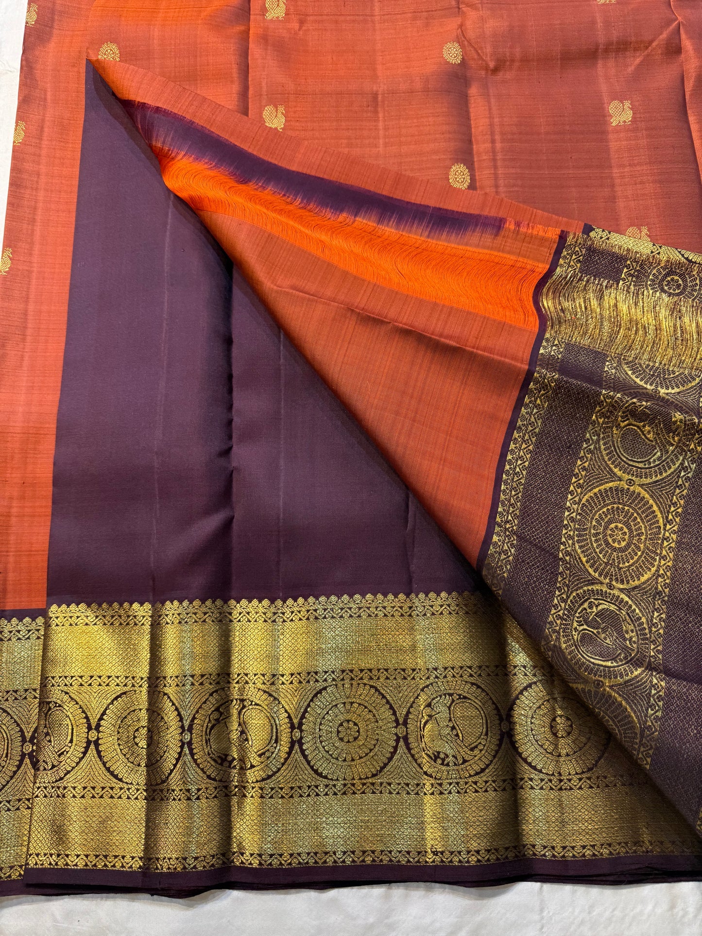 Rust Orange Supreme – Korvai Kanchipuram Pure Silk Saree
Shot rust-orange body with gold chakram & mankolam buttas all over
Deep purple korvai border with annam & chakram zari medallions
Rich vadamalli zari-brocade pallu | Contrast blouse | Fall & Pico Do