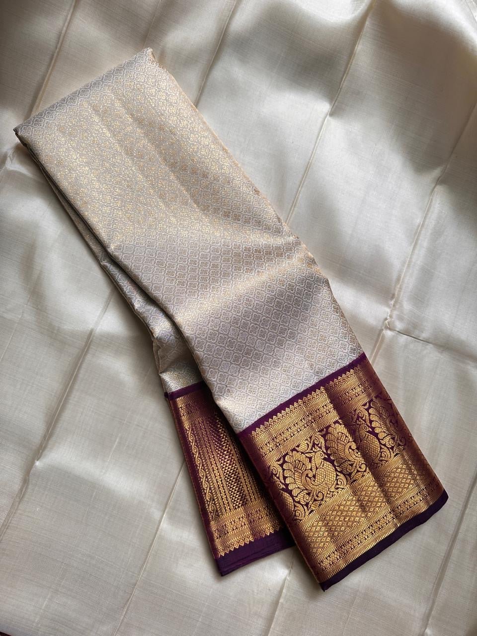 Ivory silk meets tradition in this elegant Kanchipuram saree. With a deep purple border and Mayuri motifs, it’s perfect for moments that call for quiet richness.