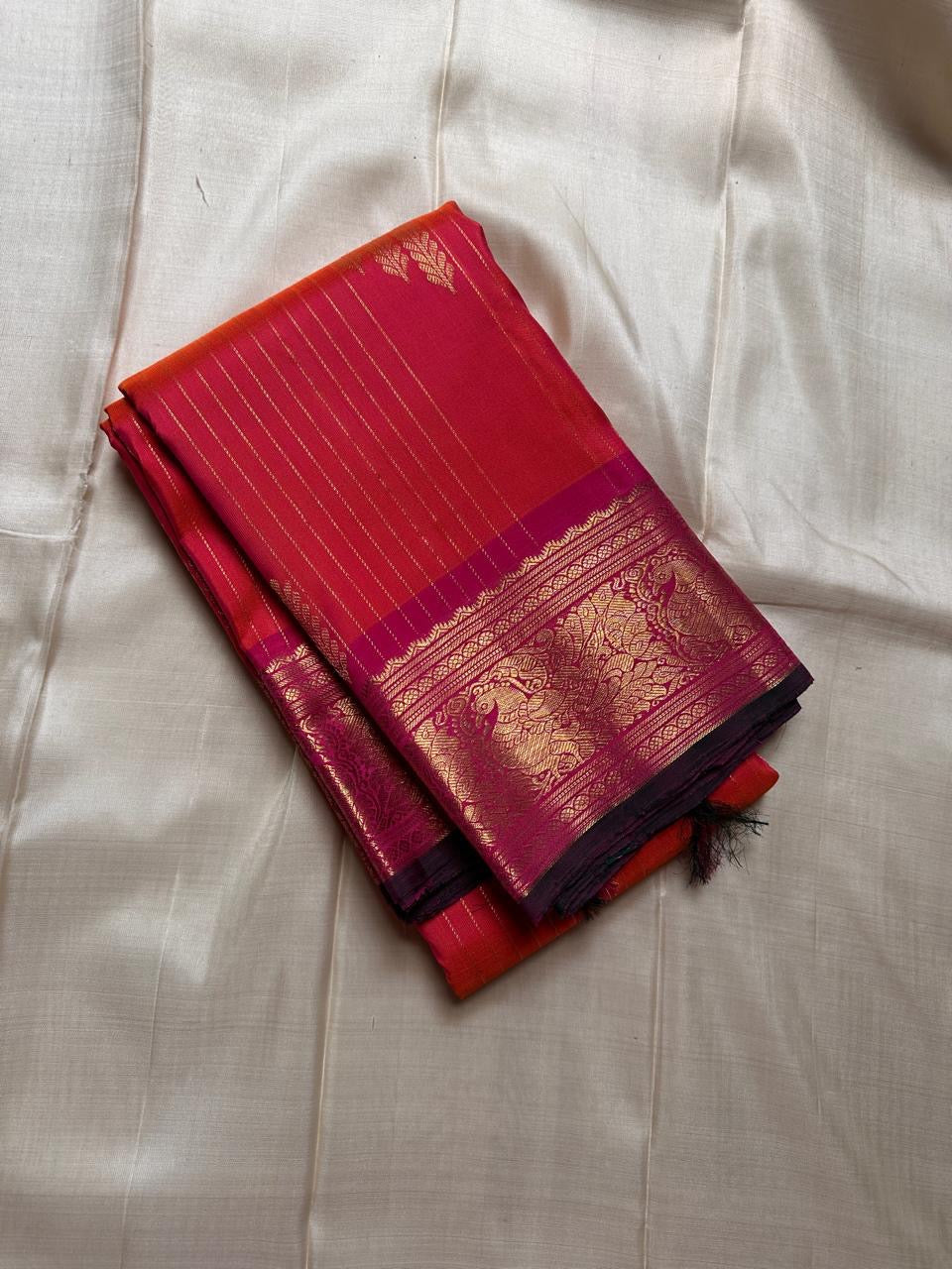 Sharvani – Orange & Magenta Pure Silk Kanchipuram Saree
Featuring zari-woven stripes across the body for subtle structure.
Bold magenta border with rice reed and peacock motifs, finished with a green selvedge.