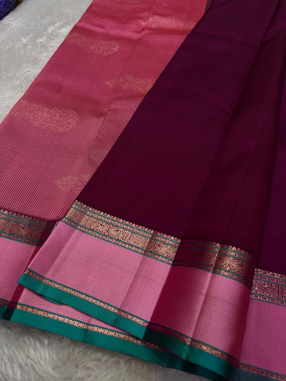 A regal magenta silk with intricate zari buttas scattered delicately across the body. The border is a stunning blend of soft pink and teal, featuring fine gold silver zari weaving and temple motifs