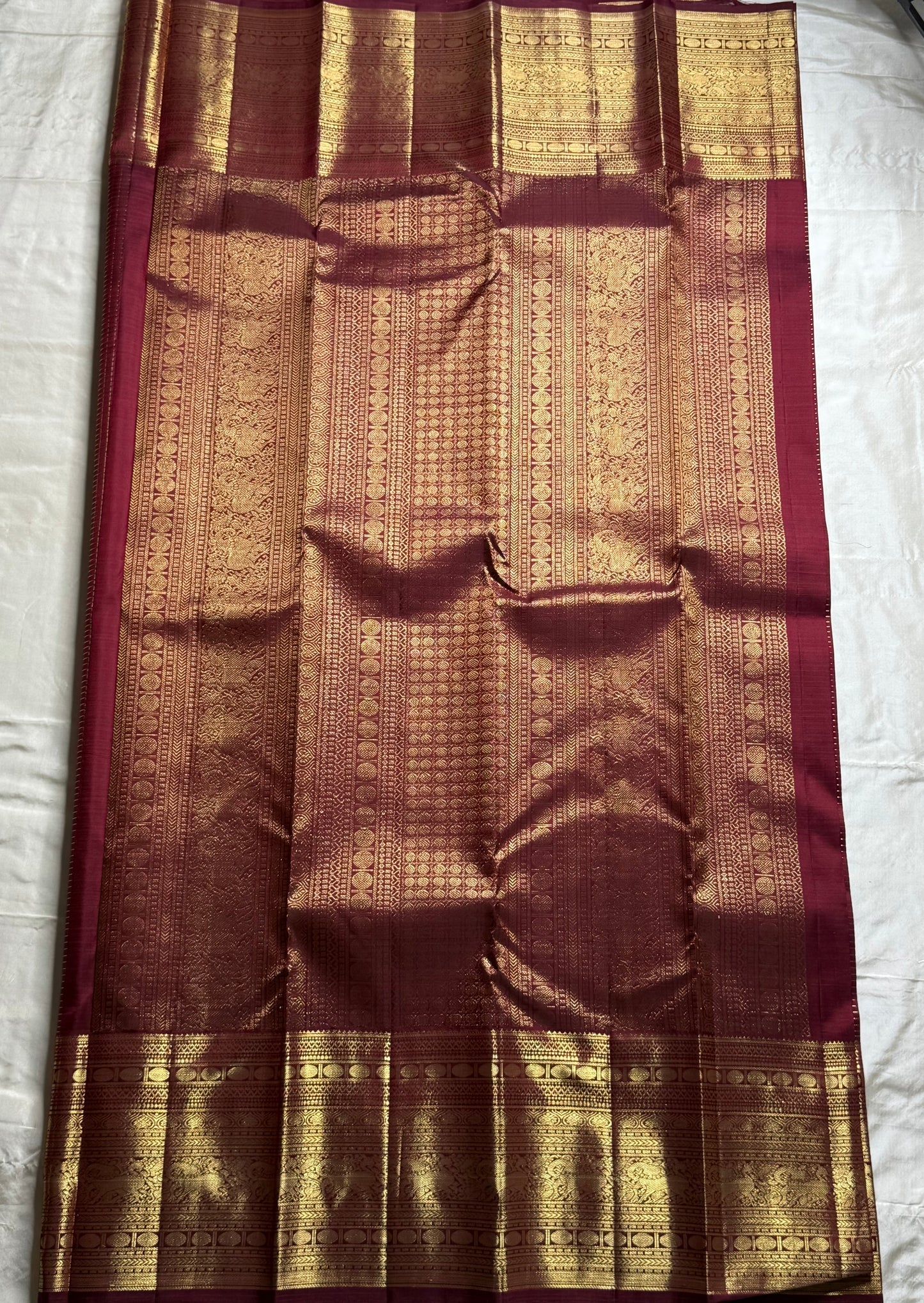 Pavila – Rich brown pure silk Kanchipuram saree in shimmering zari kattam checks. Layered varisaipet borders feature peacocks, yazhi, and rudraksham motifs in ornate zari. A bold broad yazhi adai motif lends ceremonial elegance. Grand ornate Pallu