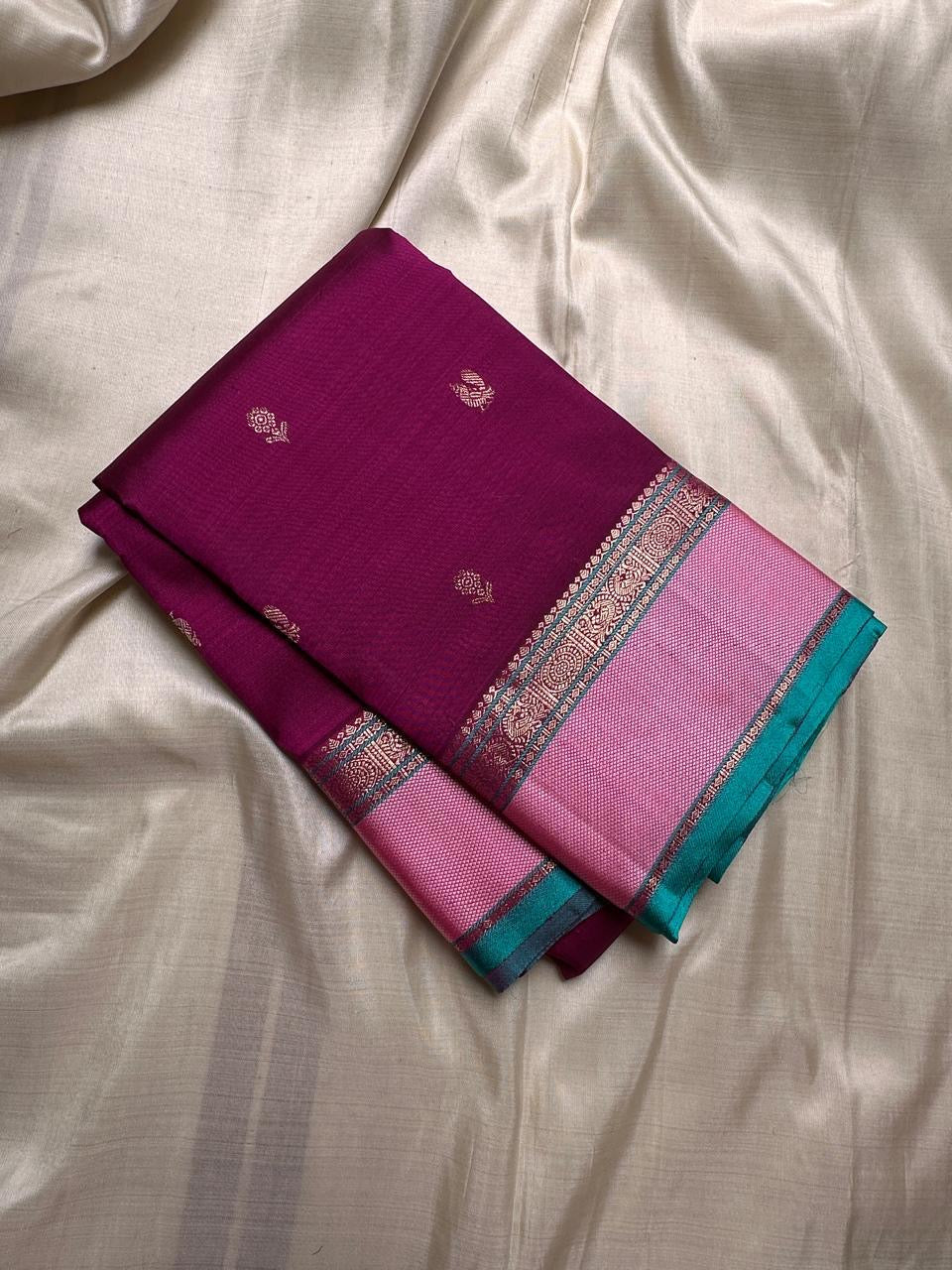 A regal magenta silk with intricate zari buttas scattered delicately across the body. The border is a stunning blend of soft pink and teal, featuring fine gold silver zari weaving and temple motifs