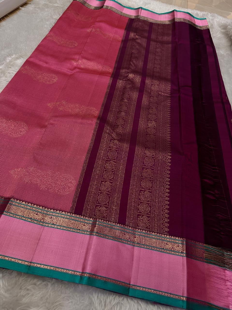 A regal magenta silk with intricate zari buttas scattered delicately across the body. The border is a stunning blend of soft pink and teal, featuring fine gold silver zari weaving and temple motifs