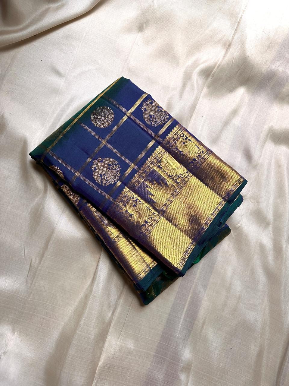 A celestial blend of blue and green, this Kanjivaram silk saree ,adorned with Gandaberunda and Kamalam buttas, golden checks, and a majestic thazhampoo Mayil border. Zari kattam