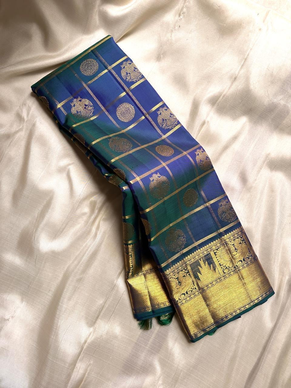 A celestial blend of blue and green, this Kanjivaram silk saree ,adorned with Gandaberunda and Kamalam buttas, golden checks, and a majestic thazhampoo Mayil border. Zari kattam