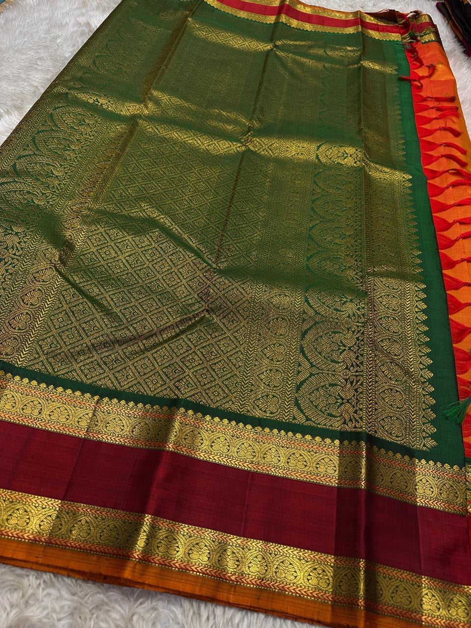 Orange Maragatha Korvai — Kanchipuram Pure Silk
Sunset aruna-orange body with delicate single zari butta
Deep maroon korvai borders with floral rosette motifs; green selvedge
Rich maragatha-green zari pallu (zari jaal & vine panels)..description
