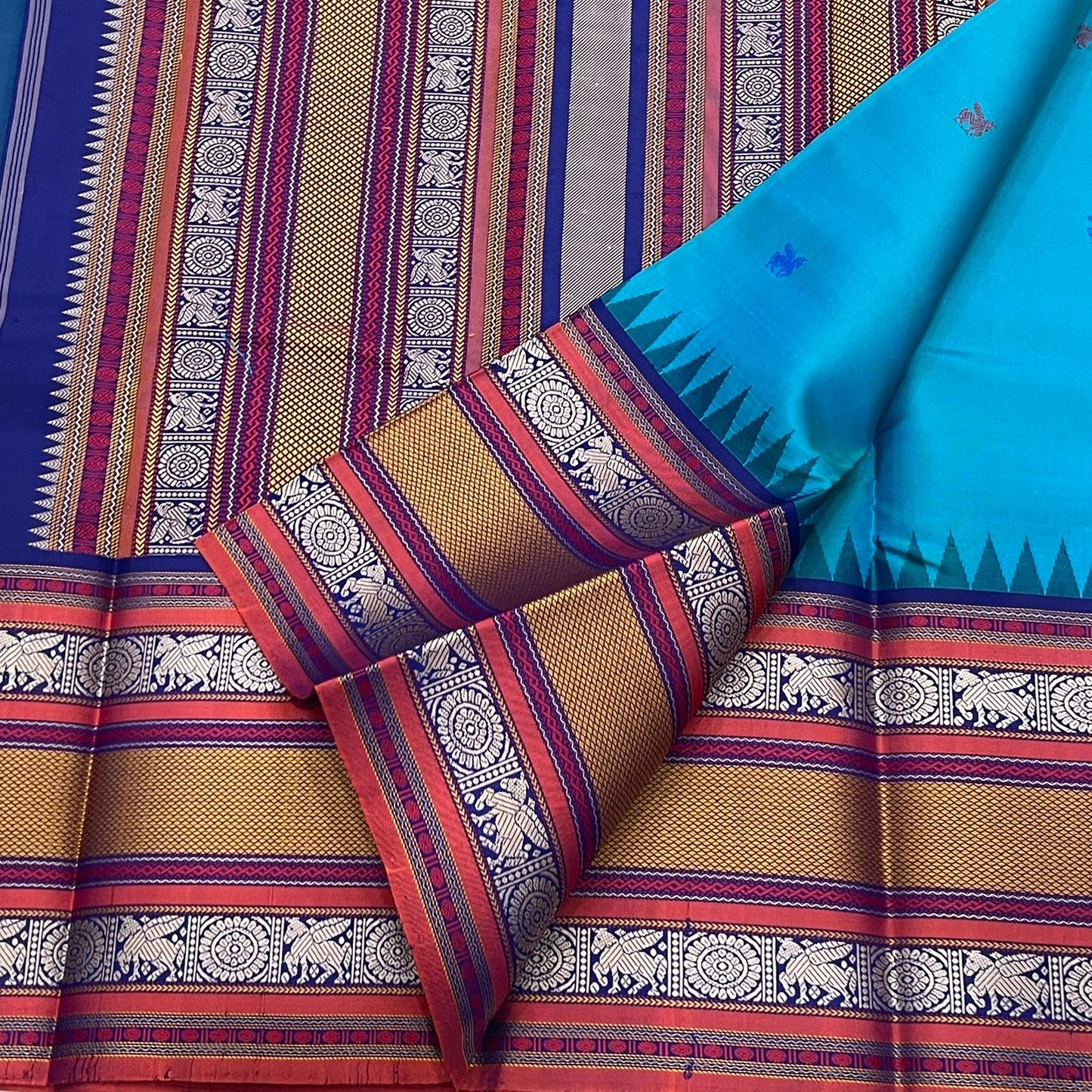 Peacock-teal body with woven silk-thread buttas in navy & coral hues
Korvai contrast ruby-maroon borders with gaja-chakram & temple motifs
Grand varisaipettu striped pallu in navy, mustard & maroon tones
Silk Mark Certified • Ships in Two Weeks