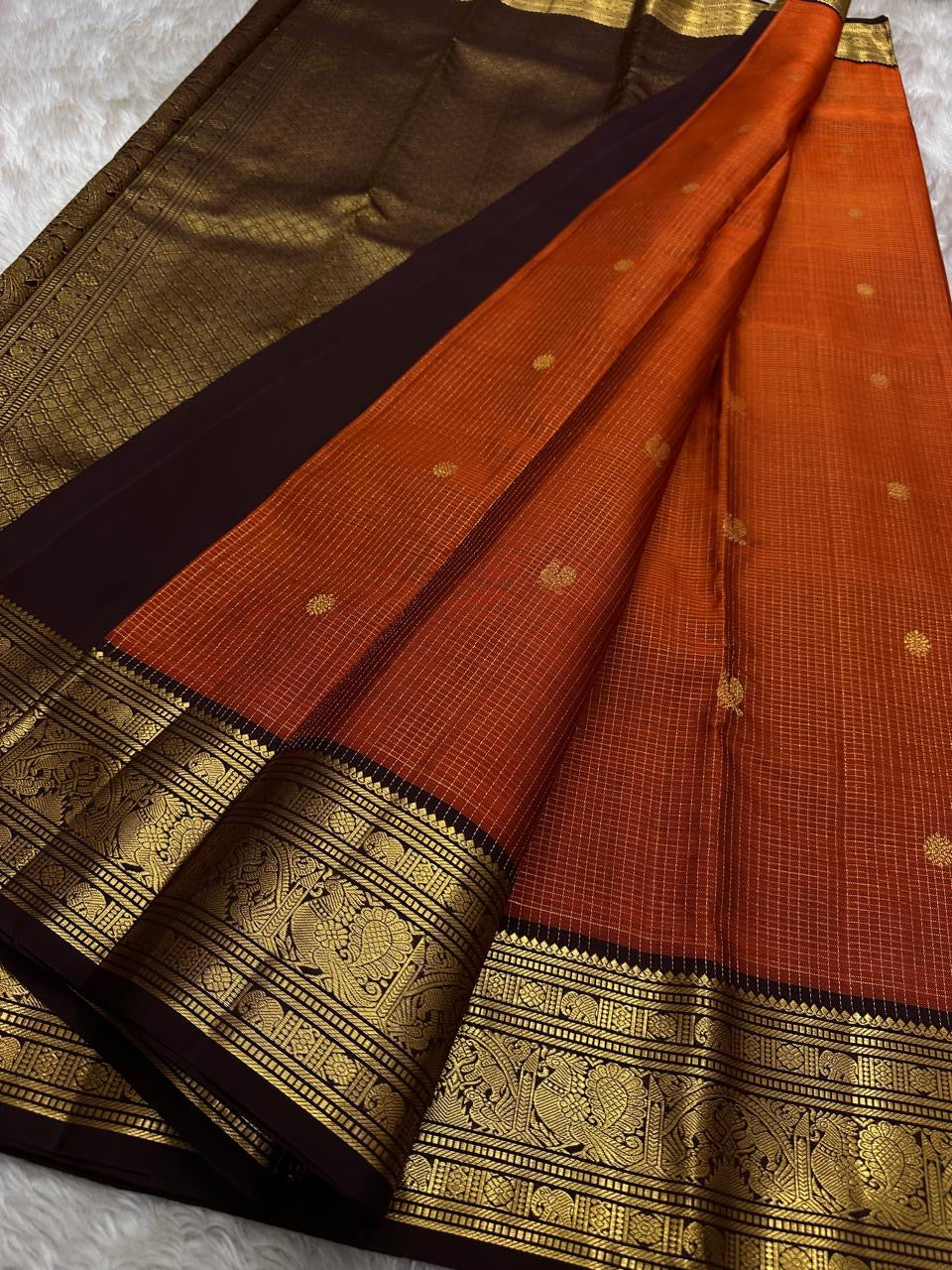 Kavi — oosi kattam Pure Kanchipuram Silk Saree
Burnt Orange (saffron–rust) body
Deep coffee-maroon contrast border with grand antique-gold zari
Rich brocade pallu; peacock & floral vines in the border
Silk Mark Certified