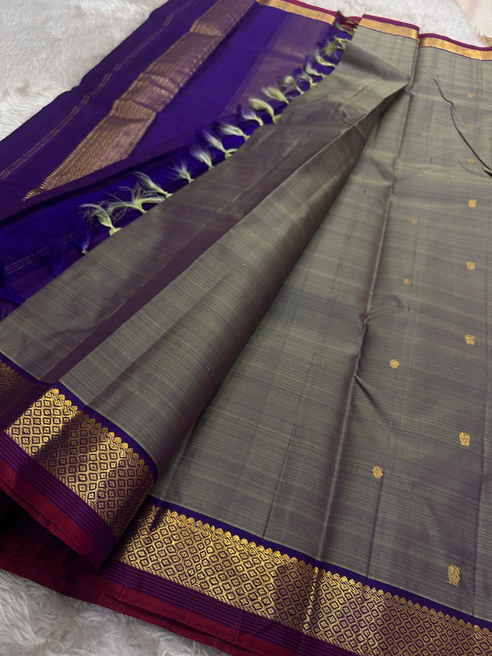 Varnam – Dove Tail Gray & Purple, a Kanchipuram silk saree where elegance meets bold expression. The soft dove tail gray base is adorned with vibrant magenta checks,Framed by a rich purple zari border and finished with an intricate contrast pallu