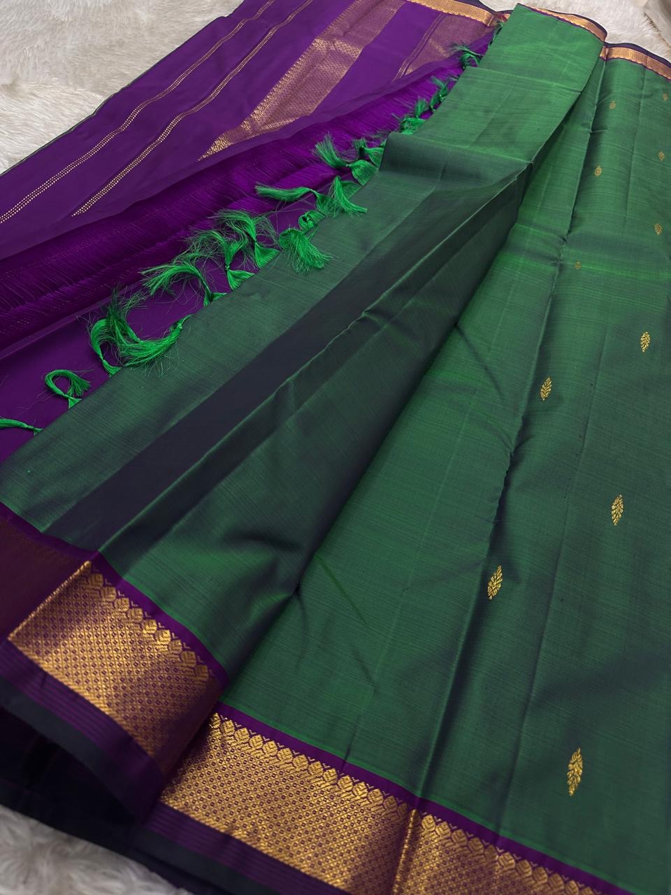 Varnam is a pure Kanchipuram silk saree in rich emerald green with subtle dual-tone shading. Woven gold zari buttas dot the body, while a petite purple border woven with intricate zari adds refined contrast. Arai maadam Pallu, jewel toned classic !