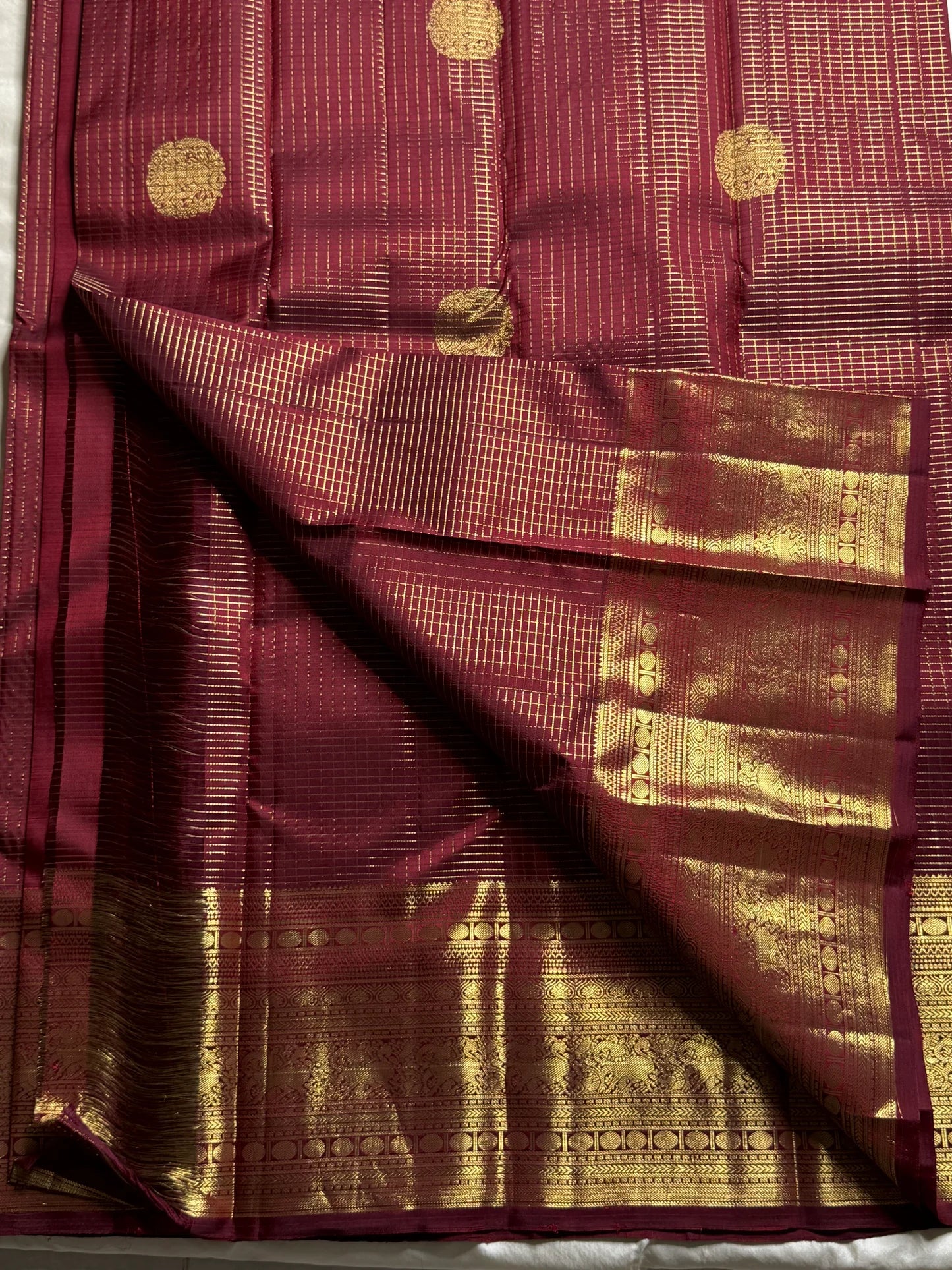 Pavila – Rich brown pure silk Kanchipuram saree in shimmering zari kattam checks. Layered varisaipet borders feature peacocks, yazhi, and rudraksham motifs in ornate zari. A bold broad yazhi adai motif lends ceremonial elegance. Grand ornate Pallu