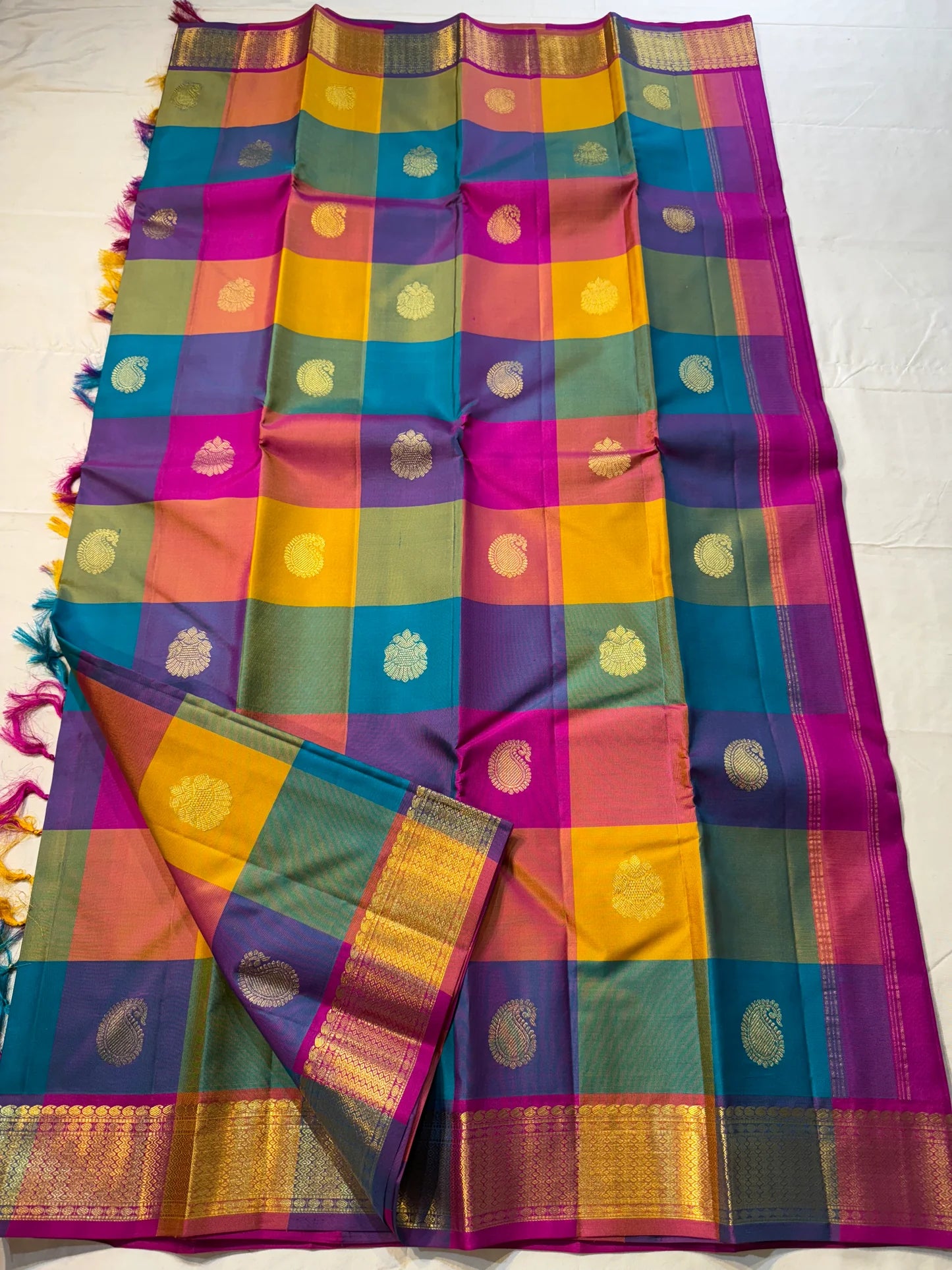 Rangoli Kattam – Pure Silk Kanchipuram Saree
Vibrant multicolor kattam checks with golden zari buttas across the body
Rich rani pink pallu with intricate zari weaving for a grand finish
Contrast purple borders edged with gleaming zari motifs