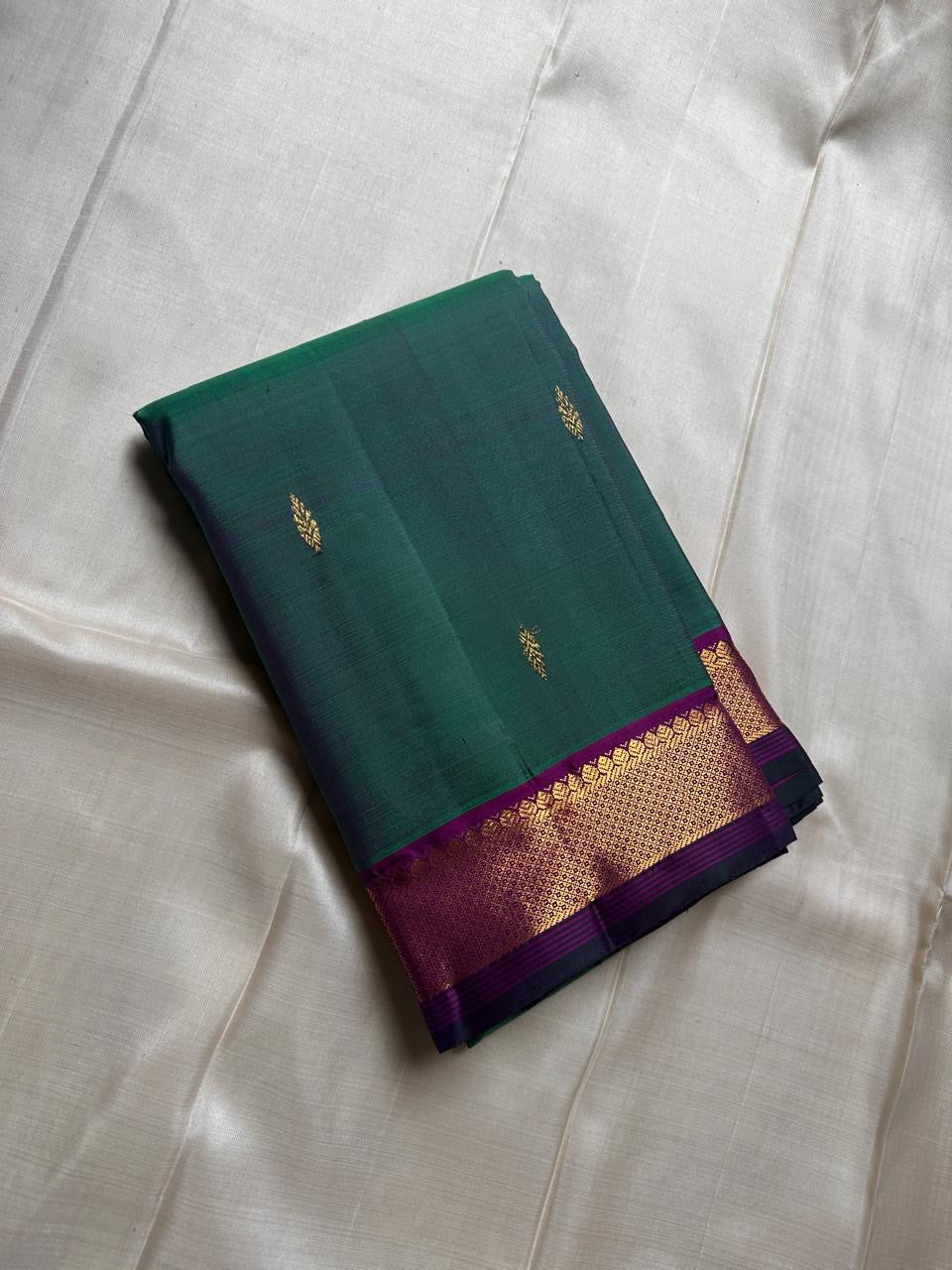 Varnam is a pure Kanchipuram silk saree in rich emerald green with subtle dual-tone shading. Woven gold zari buttas dot the body, while a petite purple border woven with intricate zari adds refined contrast. Arai maadam Pallu, jewel toned classic !