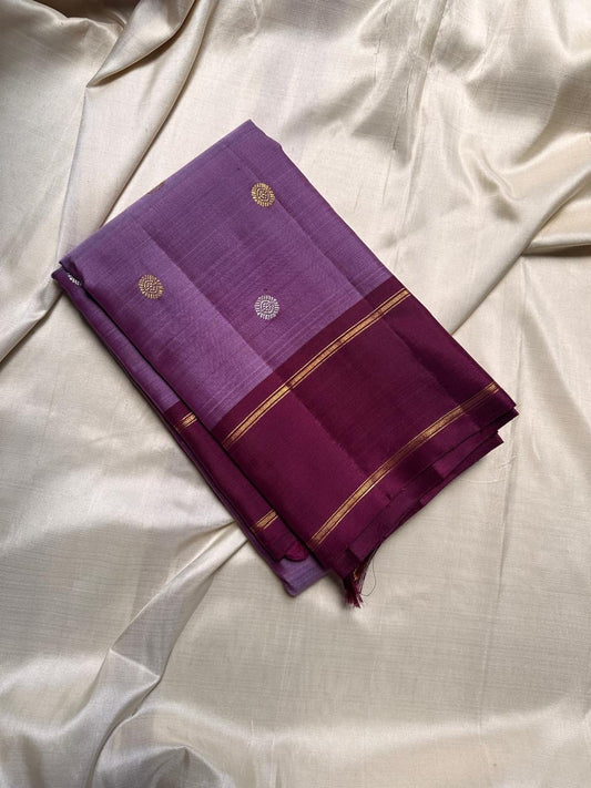 Ilam  Vadamalli Chakram — Kanchipuram Pure Silk
Pastel lilac–mauve body with gold & silver chakram buttas
Deep plum contrast border with twin kaddi zari lines
Plum pallu with elegant vertical zari stripes; lilac tassels
Silk Mark Certified •