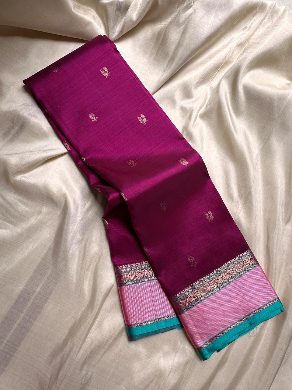A regal magenta silk with intricate zari buttas scattered delicately across the body. The border is a stunning blend of soft pink and teal, featuring fine gold silver zari weaving and temple motifs