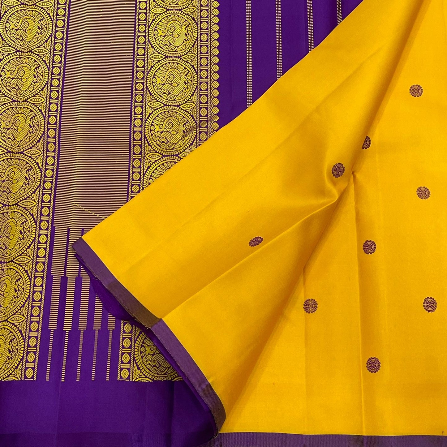 Manjal Vadamalli Chakram – Pure Kanchipuram Silk Saree
Marigold yellow body with purple floral thread buttas
Grand royal purple pallu with gold medallion zari & reku-style stripes
Handwoven Double Warp Weave | Silk Mark Certified | Fall & Pico Done