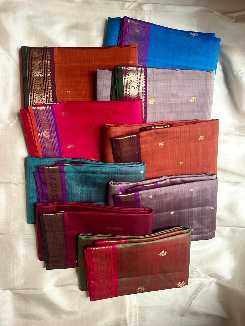 Assorted folded pure Kanchipuram silk sarees in multiple colors arranged together on a light fabric backdrop.