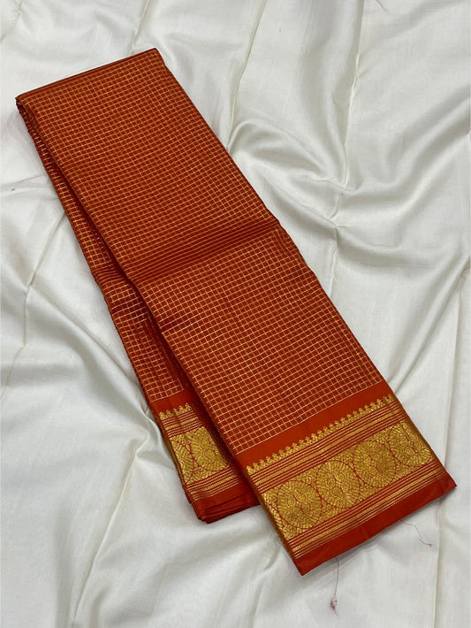 Folded burnt orange Kanchipuram silk saree on white backdrop with antique gold zari woven border and fine check body