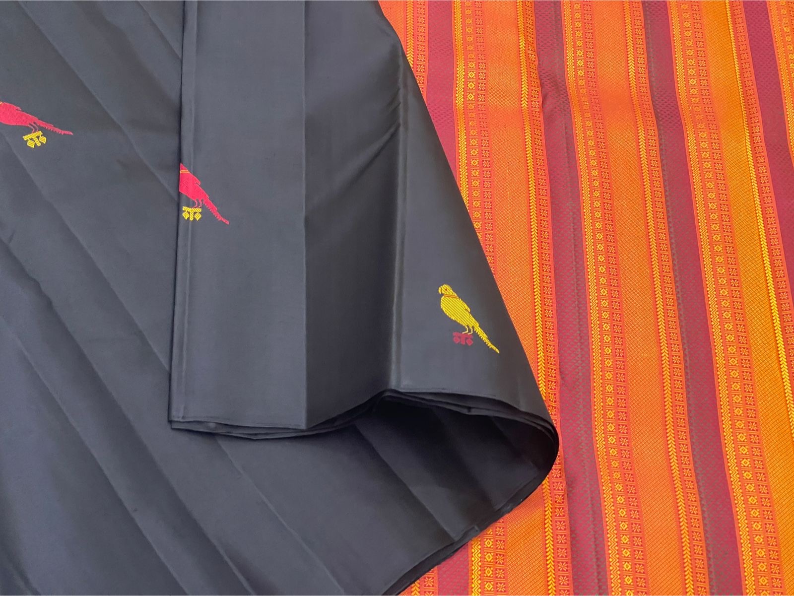 Black Pure Kanchipuram Silk Saree fabric with a yellow bird motif draped beside an orange striped pallu with pink border.