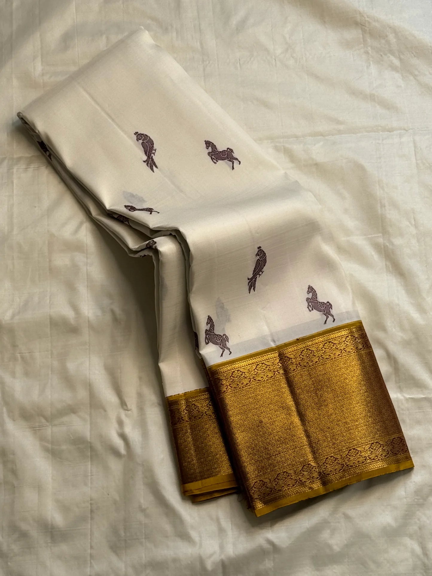 Folded ivory Kanchipuram silk saree with maroon horse and parrot buttas and rich antique-gold zari border on cream backdrop