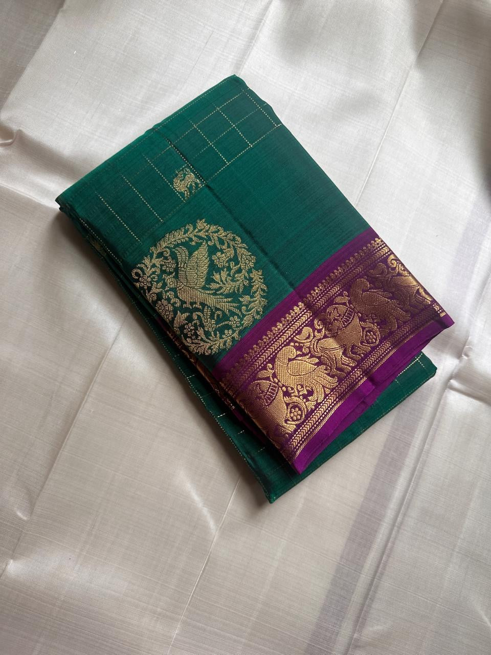 Sorgavaasal Kili – Pure Silk Kanchipuram Saree in Teal with Purple Zari Kattam
Intricately handwoven with traditional zari kattam checks and adorned with exquisite zari-woven Yazhi buttas, this saree features a vibrant teal body paired with a regal purple