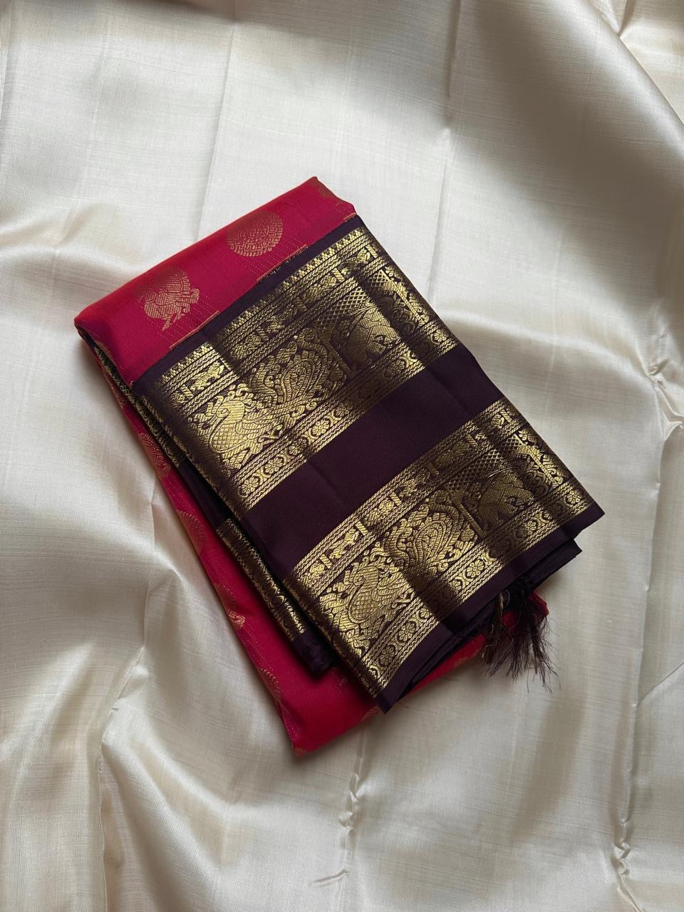 A regal  shot tomato red pink Kanchipuram silk saree features elegant chakram and mayil gold zari buttas woven across the body. Brown contrast rettapet border and Pallu vanasringaram details