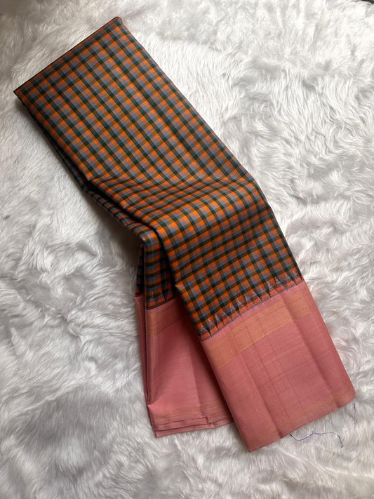 Nirai – A Madras-Inspired Kanchipuram Silk in Amber & Pastel Pink.
Woven with fine podi kattam checks in black, amber orange, and grey.
The pastel pink border with pillayar moggu detailing adds softness, with a  seer zari pallu