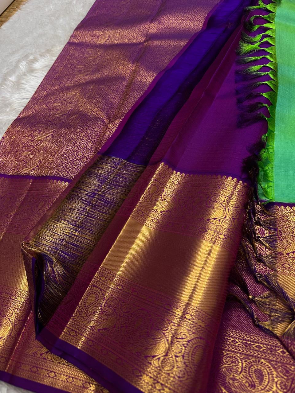 This pure silk Kanchipuram saree features a radiant shot color blend of green, gold, and blue, adorned with delicate zari buttas. A regal royal purple rettapet border and an elaborate peacock motif pallu complete this timeless drape — perfect for festive