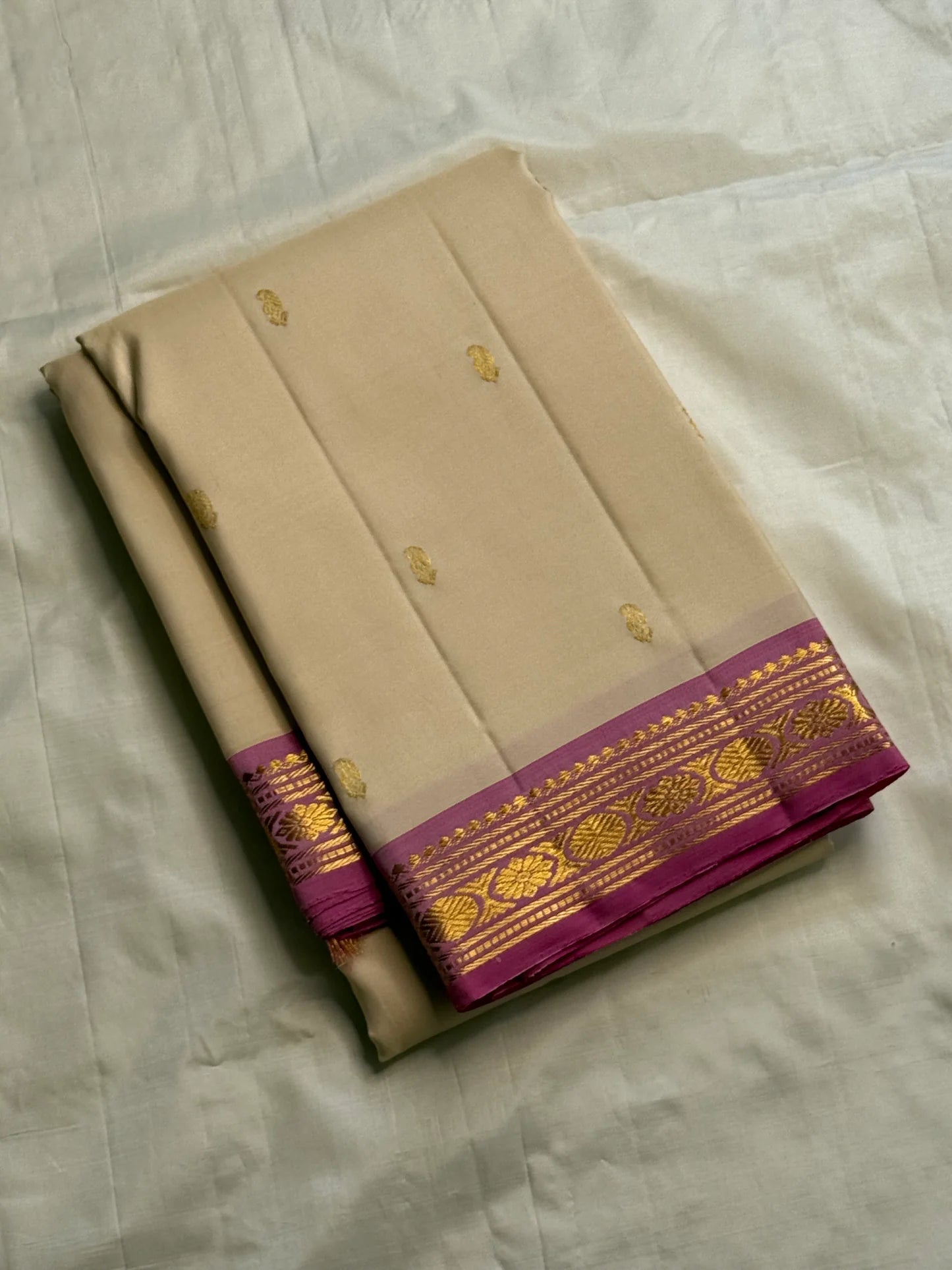 Folded cream silk saree with small gold buttas and a magenta border featuring gold zari woven motifs, on white cloth.
