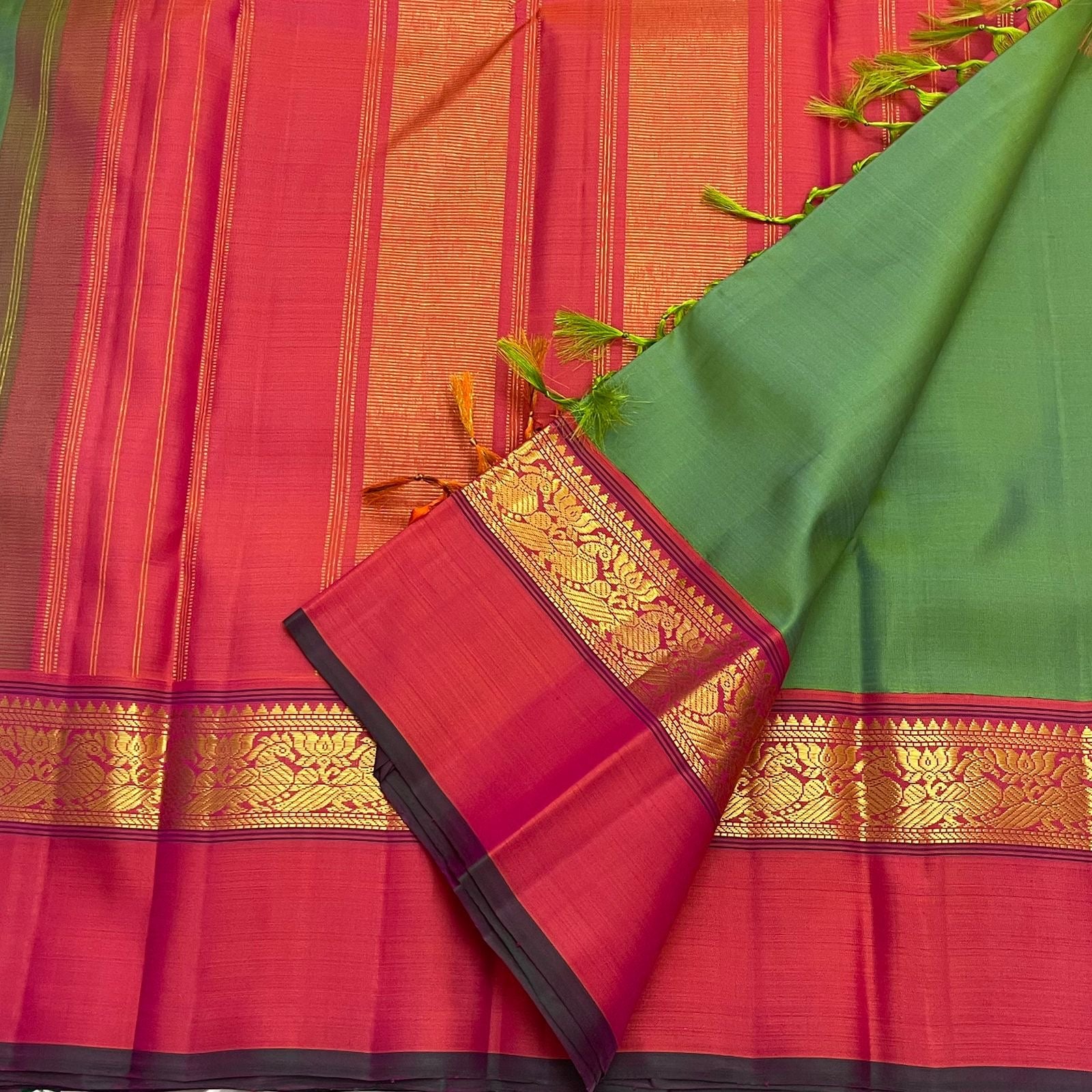 Green saree with pink gold zari border placed over a pink pallu with vertical zari stripes and green tassels.