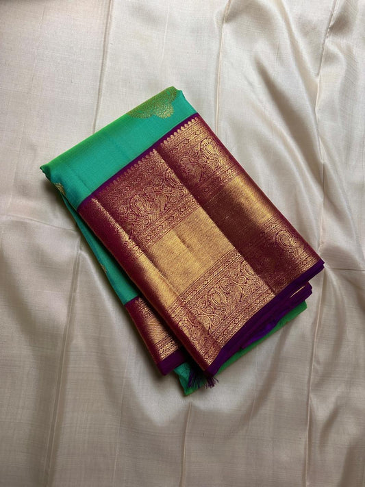 This pure silk Kanchipuram saree features a radiant shot color blend of green, gold, and blue, adorned with delicate zari buttas. A regal royal purple rettapet border and an elaborate peacock motif pallu complete this timeless drape — perfect for festive