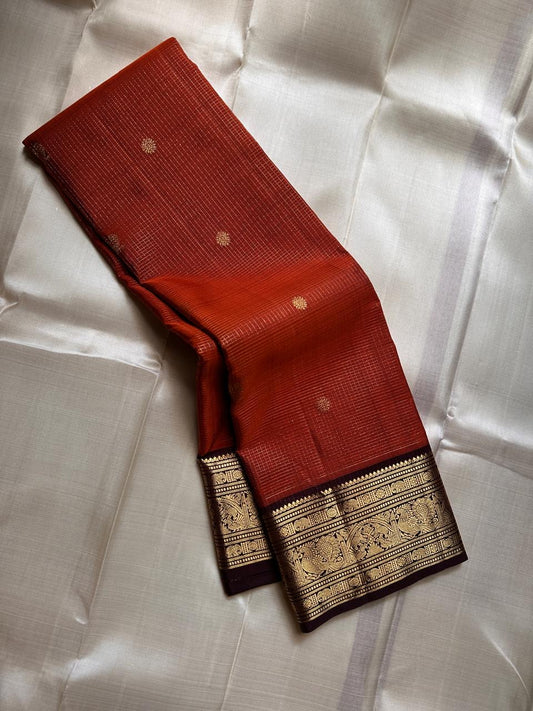 Kavi — oosi  kattam Pure Kanchipuram Silk Saree

Burnt Orange (saffron–rust) body
Deep coffee-maroon contrast border with grand antique-gold zari
Rich brocade pallu; peacock & floral vines in the border
Silk Mark Certified