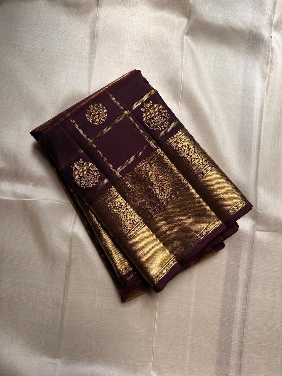 Folded plum Kanchipuram silk saree with Gandaberunda zari kattam checks and gold elephant border on an ivory silk background.